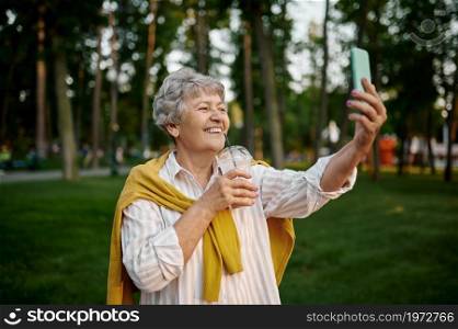 Smiling granny makes selfie with milk coctail in summer amusement park, back to childhood. Aged people lifestyle. Funny grandmother having fun outdoors, old female person on nature. Smiling granny makes selfie with milk coctail