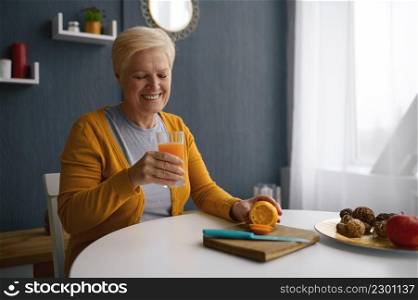 Smiling grey-haired woman holding glass cup with orange juice. Freshly picked oranges on table. Smiling grey-haired woman holding glass with juice