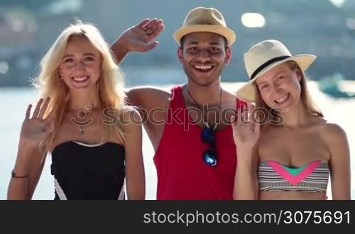 Smiling group of interracial people wearing swimsuits waving hello with hands on the beach during summer vacation at seaside.