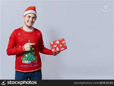 Smiling guy in christmas hat holding and pointing a gift box. Portrait of teenager in christmas hat holding and pointing a gift box. Guy in christmas hat and sweater holding a gift box isolated
