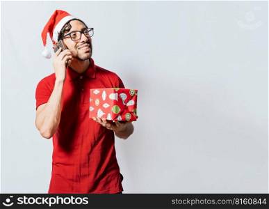 Smiling guy in christmas hat holding christmas gift box and talking on the phone. Christmas man holding gift and calling family on phone. Happy man holding gift and calling family at christmas