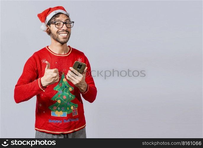 Smiling guy in christmas hat using cellphone with thumb up. Smiling young man in christmas hat using cellphone and giving thumbs up. People in santa hat using phone and doing ok gesture