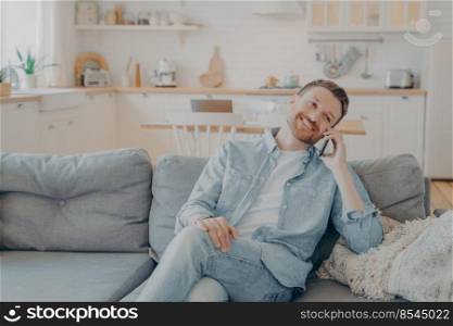 Smiling guy sits on couch at home having fun while talking on modern cellphone gadget, happy young male relaxing on sofa in living room while making conversation with his girlfriend. Smiling guy sits on couch at home having fun while talking on modern cellphone gadget