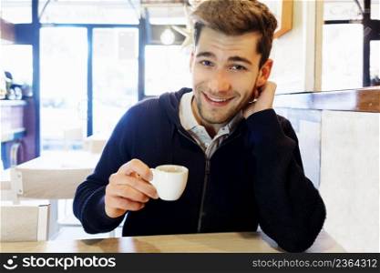 Smiling handsome barista sitting with coffee at the bar of the modern cafe interior
