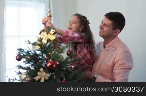 Smiling handsome father holding his adorable cheerful daughter in his arms while family decorating Christmas tree top together at home. Cute little girl putting on main decorative toy on xmas tree while her affectionate dad holding her in his arms.
