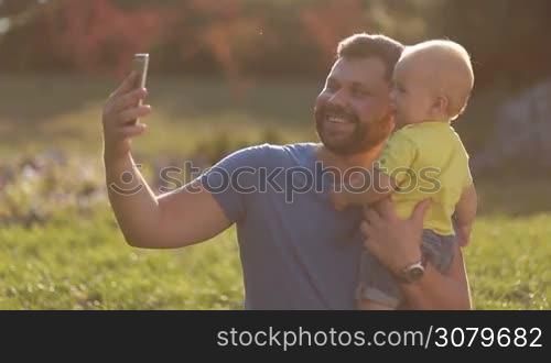 Smiling handsome father taking selfie with smartphone with his infant child as they spend leisure in summer park. Positive hipster dad carrying his cute toddler son in his hands and making self portrait on mobile phone outdoors.