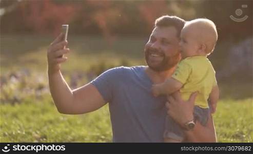 Smiling handsome father taking selfie with smartphone with his infant child as they spend leisure in summer park. Positive hipster dad carrying his cute toddler son in his hands and making self portrait on mobile phone outdoors.