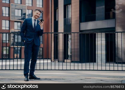 Smiling handsome male office employee dressed in blue suit checking his phone while having break, going through messages and notifications, standing alone outside with modern office building behind. Male office worker chatting on smartphone while work break outdoors