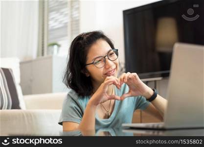 Smiling Happy Asian woman in blue shirt and eyeglasses making her hands in heart shape during video call with people and her family via internet technology on laptop computer