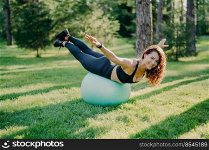 Smiling happy brunette woman with european appearance balances on fitness ball stretches arms dressed in top and leggings poses outdoor in summer park doing pilates at nature during sunny day