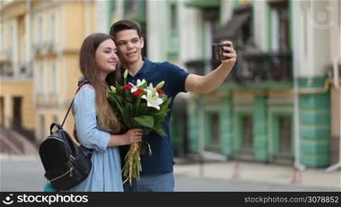 Smiling happy couple in love taking selfie with mobile phone during romantic date outdoors. Attractive brunette woman holding bunch of beautiful flowers embracing her handsome boyfriend while couple making self portrait with smartphone.