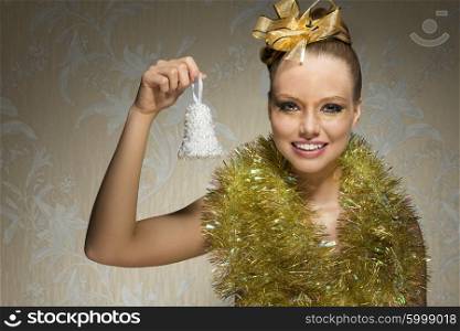 smiling happy female with freckles and artistic christmas look. Posing with shiny tinsel around neck, golden ribbon in the hair-style and christmas glossy make-up. Taking little bell in the hand
