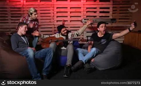 Smiling happy teenage musicians taking selfie photo with mobile phone while sitting on bean bag chairs after rehearsal in the club. Group of cheerful young men making selfie using cellphone over musical instruments background.