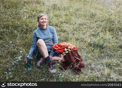 Smiling happy woman with backpack resting on grass during a hike in the mountains, actively spending summer vacation
