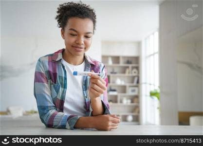 Smiling healthy mixed race girl holding digital thermometer, pleased with normal body temperature after treatment, sitting at home. Happy young woman checking temp during coronavirus pandemic.. Smiling healthy mixed race girl holding thermometer pleased with normal body temperature indoors