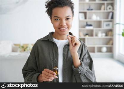 Smiling healthy young mixed race girl holding pill, medicine jar, standing at home. Happy pretty lady takes daily medication, vitamins, dietary supplement for beauty, skin, hair. Health care, wellness. Smiling healthy young mixed race girl takes daily medicine pills, vitamins, supplements for beauty