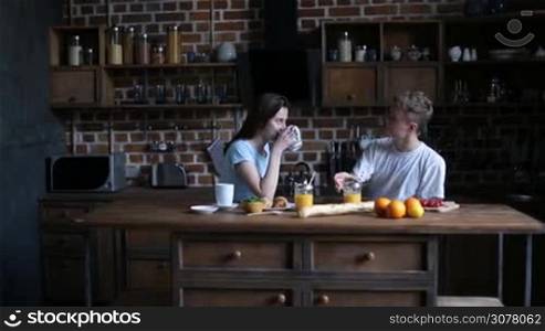 Smiling hipster couple spending leisure, communicating while sitting at the table and having breakfast in domestic kitchen in the morning. Cheerful man telling story while his cute girlfriend drinking coffee.