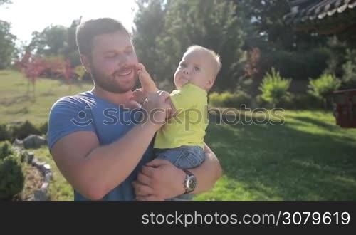 Smiling hipster father holding his cute toddler son and kissing with love and tenderness while walking and enjoying time together in summer park. Stylish dad and his little infant baby boy bonding with each other outdoors in rays of setting sun.