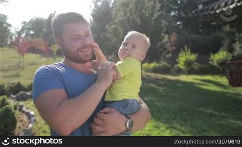 Smiling hipster father holding his cute toddler son and kissing with love and tenderness while walking and enjoying time together in summer park. Stylish dad and his little infant baby boy bonding with each other outdoors in rays of setting sun.