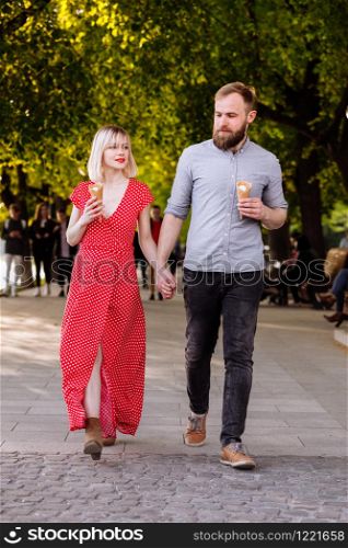 smiling hipsters couple eating ice cream and having fun in the city. stylish young man with beard and blonde woman in red dress are walking in the street. smiling hipsters couple eating ice cream and having fun in the city. stylish young man with beard and blonde woman in red dress are walking in the street.