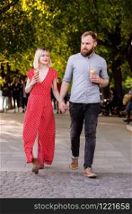 smiling hipsters couple eating ice cream and having fun in the city. stylish young man with beard and blonde woman in red dress are walking in the street. smiling hipsters couple eating ice cream and having fun in the city. stylish young man with beard and blonde woman in red dress are walking in the street.