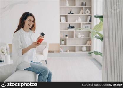 Smiling hispanic female holding bank credit card, smartphone, making successful payment, shopping in online store, sitting in living room at home. E commerce, e-bank apps, banking services advertising. Online banking. Happy female holding bank credit card, smartphone, makes cashless payment at home
