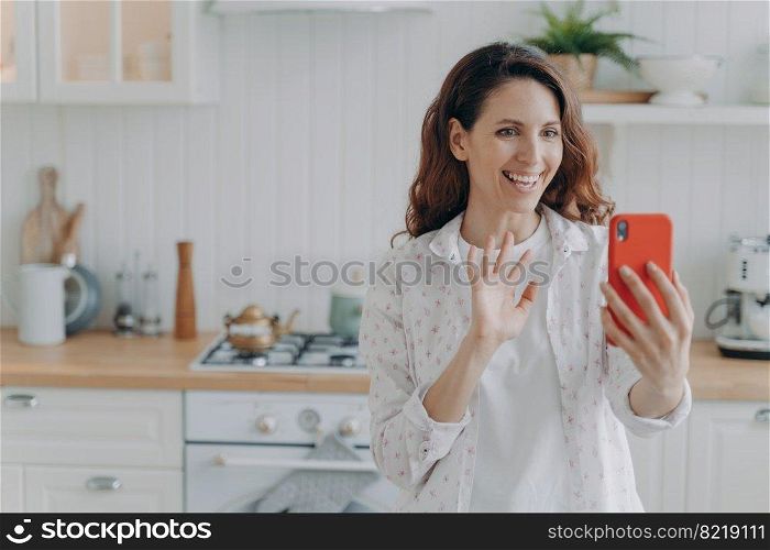 Smiling hispanic female holding smartphone talks by videocall waving hand to camera. Positive woman calling, using phone app, greeting friend, enjoying remote communication in kitchen at home.. Smiling girl holding phone talk by videocall waving hand to camera in kitchen. Remote communication