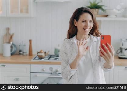 Smiling hispanic female holding smartphone talks by videocall waving hand to camera. Positive woman calling, using phone app, greeting friend, enjoying remote communication in kitchen at home.. Smiling girl holding phone talk by videocall waving hand to camera in kitchen. Remote communication