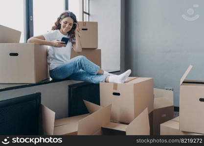 Smiling hispanic female sit on windowsill with carton boxes use smartphone apps buy modern furniture for first own new house or search moving company, arranging relocation. Ecommerce services, removal. Smiling girl use phone apps buy furniture for new home, search moving company. Ecommerce, relocation