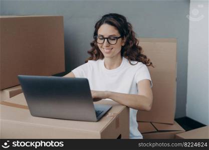 Smiling hispanic female wearing glasses working at laptop surrounded with cardboard boxes, preparing customers orders, parcels of online store for sending. Delivery service, e-commerce concept.. Girl works at laptop preparing parcels of online store for sending. Delivery service, ecommerce