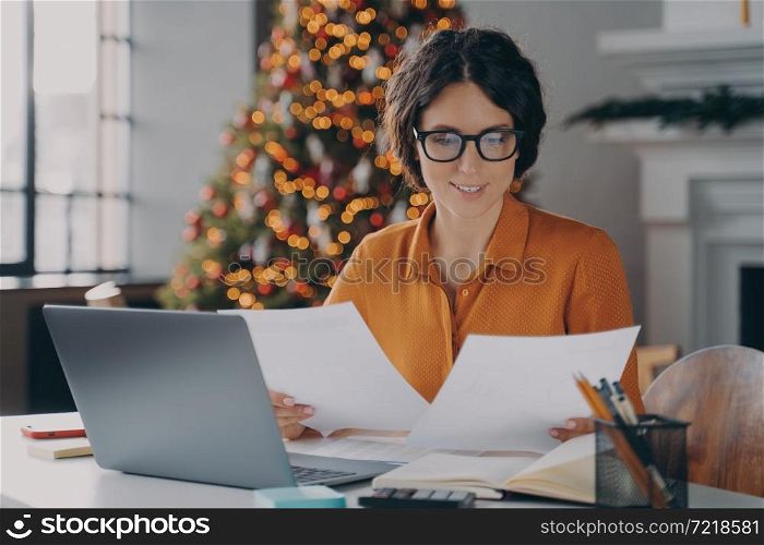 Smiling Hispanic woman employee working in office at christmas time, female checking invoice documents and company reports for year-end financial meeting sits at desk with laptop against New Year tree. Smiling Hispanic woman employee working in office during christmas time