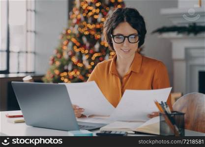 Smiling Hispanic woman employee working in office at christmas time, female checking invoice documents and company reports for year-end financial meeting sits at desk with laptop against New Year tree. Smiling Hispanic woman employee working in office during christmas time