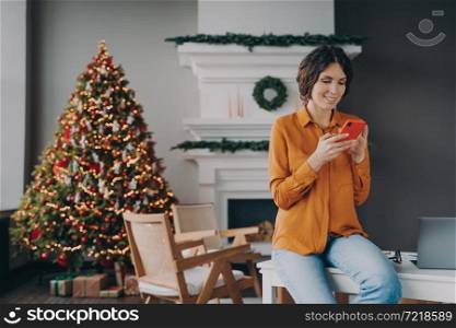 Smiling hispanic woman in casual clothes reading message on mobile phone during remote work at home on Christmas holidays, pleased female using smartphone while sitting in room decorated for xmas. Smiling hispanic woman using mobile phone during remote work at home on Christmas holidays