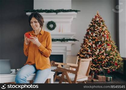 Smiling hispanic woman in casual clothes reading message on mobile phone during remote work at home on Christmas holidays, pleased female using smartphone while sitting in room decorated for xmas. Smiling hispanic woman using mobile phone during remote work at home on Christmas holidays