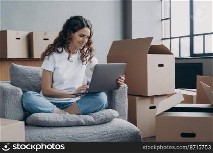 Smiling hispanic woman using laptop, looking at screen, sitting in armchair in living room with cardboard boxes with belongings on relocation day. Female choosing moving service for moving in new home. Smiling hispanic woman using laptop choosing moving service for moving in new home on relocation day