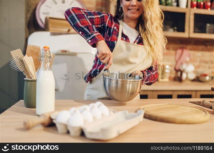 Smiling housewife in an apron making dough in bowl, kitchen interior on background. Female cook prepares fresh homemade cake. Domestic pie preparation. Smiling housewife in apron making dough in bowl