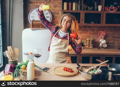 Smiling housewife in headphones and apron holding fresh pepper in hands and dance on the kitchen. Female kitchener cooking healthy vegetarian food, salad preparation. Housewife in headphones and apron dance on kitchen