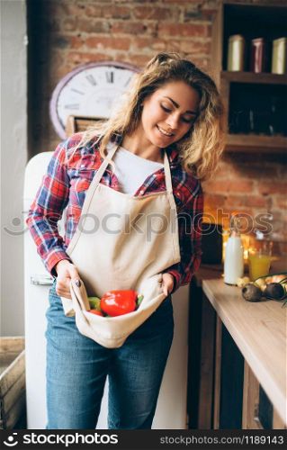 Smiling housewife keeps fresh vegetables in an apron, kitchen interior on background. Female cook making healthy vegetarian food, salad preparation. Smiling housewife keeps fresh vegetables in apron