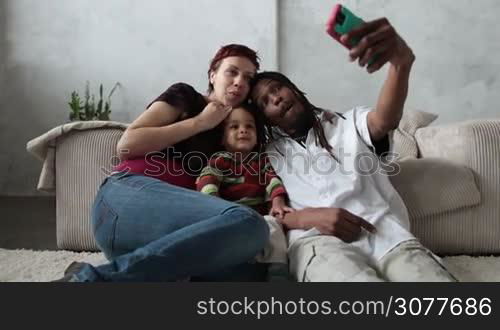 Smiling interracial family with mixed race toddler son sitting on the floor in the living room and taking selfie with smart phone. Mixed race parents and sweet child making self portrait with cellphone at home. Slow motion.