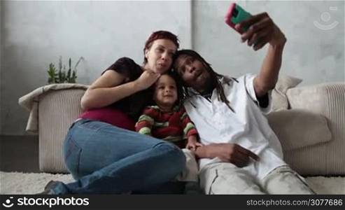 Smiling interracial family with mixed race toddler son sitting on the floor in the living room and taking selfie with smart phone. Mixed race parents and sweet child making self portrait with cellphone at home. Slow motion.