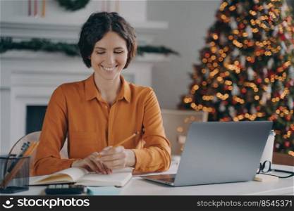 Smiling italian female making notes, using laptop at home office during christmas holidays, working online remotely in room with decorated xmas tree, writing shopping list for New Year celebration. Smiling italian female making notes, using laptop at home office during christmas holidays