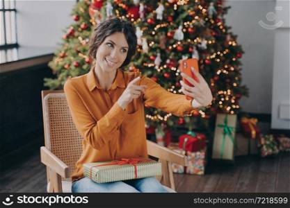 Smiling Italian woman online blogger making selfie in living room on background of decorated Xmas tree with many gifts, happy female shares happy moment of winter Christmas holidays with followers. Happy Italian female blogger shares happy moment of winter Christmas holidays with followers