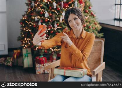 Smiling Italian woman online blogger making selfie in living room on background of decorated Xmas tree with many gifts, happy female shares happy moment of winter Christmas holidays with followers. Happy Italian female blogger shares happy moment of winter Christmas holidays with followers