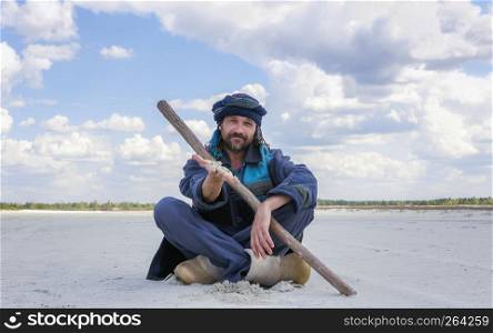 Smiling kind man in a turban and ethnic clothes demonstrates white sand by hand sitting in a desert area under a blue sky on summer day. Selective focus, conceptual image with space for copy.