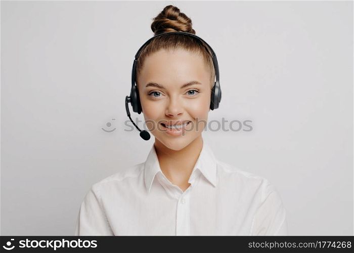 Smiling knowledgeable female call center worker in white shirt with hair in bun and black headset happy to serve and help customers, standing alone in front of grey background. Female call center worker in white shirt smiling at camera