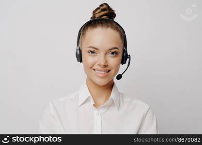 Smiling knowledgeable female call center worker in white shirt with hair in bun and black headset happy to serve and help customers, standing alone in front of grey background. Female call center worker in white shirt smiling at camera