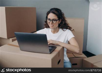 Smiling latina woman in glasses works at laptop surrounded by cardboard boxes with things on relocation day. Pleased female homeowner renter searching moving company, shopping online for new home.. Smiling female homeowner renter searching moving company at laptop, shopping online for new home