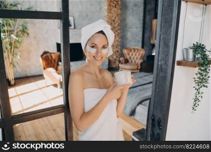 Smiling latina woman with under eye moisturizing patches holding cup of morning coffee looking at camera. Attractive girl wearing white towel enjoying skincare procedure after shower.. Smiling girl with under eye patches hold cup of morning coffee enjoy skincare procedure after shower