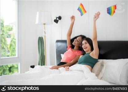 Smiling lesbian women with rainbow flags sitting on bed in bedroom at home