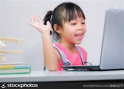 Smiling little Asian girl studying online having video call distant class with teacher using laptop. Happy little girl wave greeting with tutor while studying online with laptop at home. homeschooling